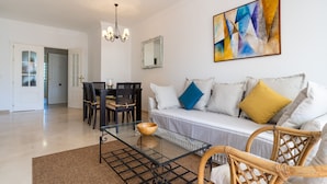 A living room with a glass coffee table, a sofa with cushions, a dining table with chairs, a chandelier, and a painting on the wall.