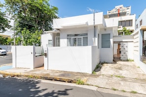 Un edificio moderno blanco con entrada protegida por una reja, un pequeño jardín y una puerta de garaje visible.