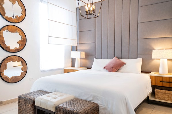 A modern bedroom with a large bed, two bedside tables with lamps, a wall-mounted mirror, and a woven bench.