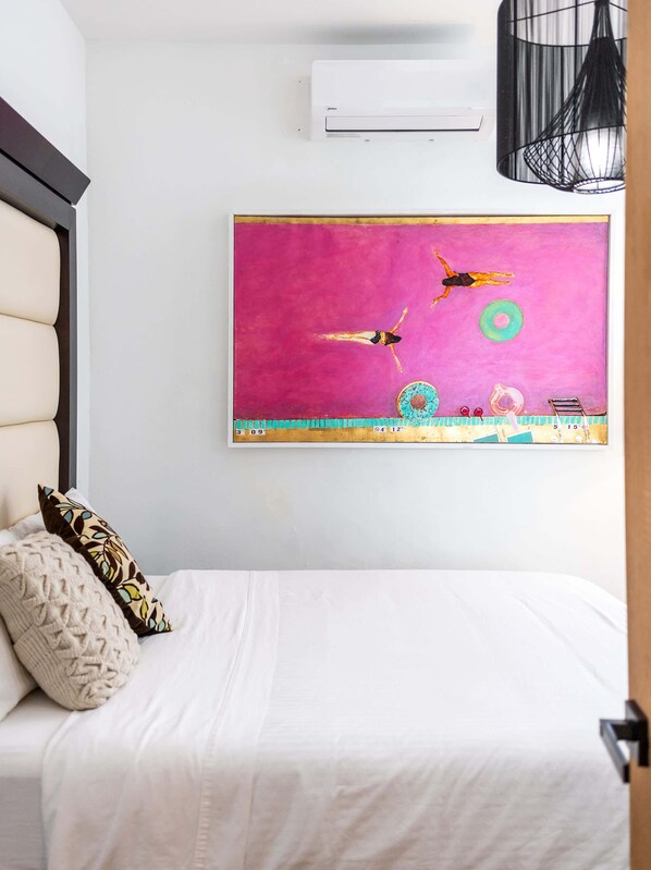 A bedroom with a bed, pillows, a wall-mounted air conditioner, and a colorful painting.