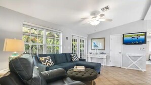 Ein Wohnzimmer mit einem dunkelgrünen Sofa, einem kleinen Hocker, einem Deckenventilator und einem an der Wand befestigten Fernseher.