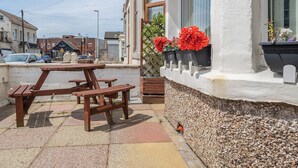 An outdoor patio with a wooden table and benches, a white building with a window displaying red flowers, and a street with cars and buildings in the background. - 7 bedroom accommodation in Blackpool (Blackpool)