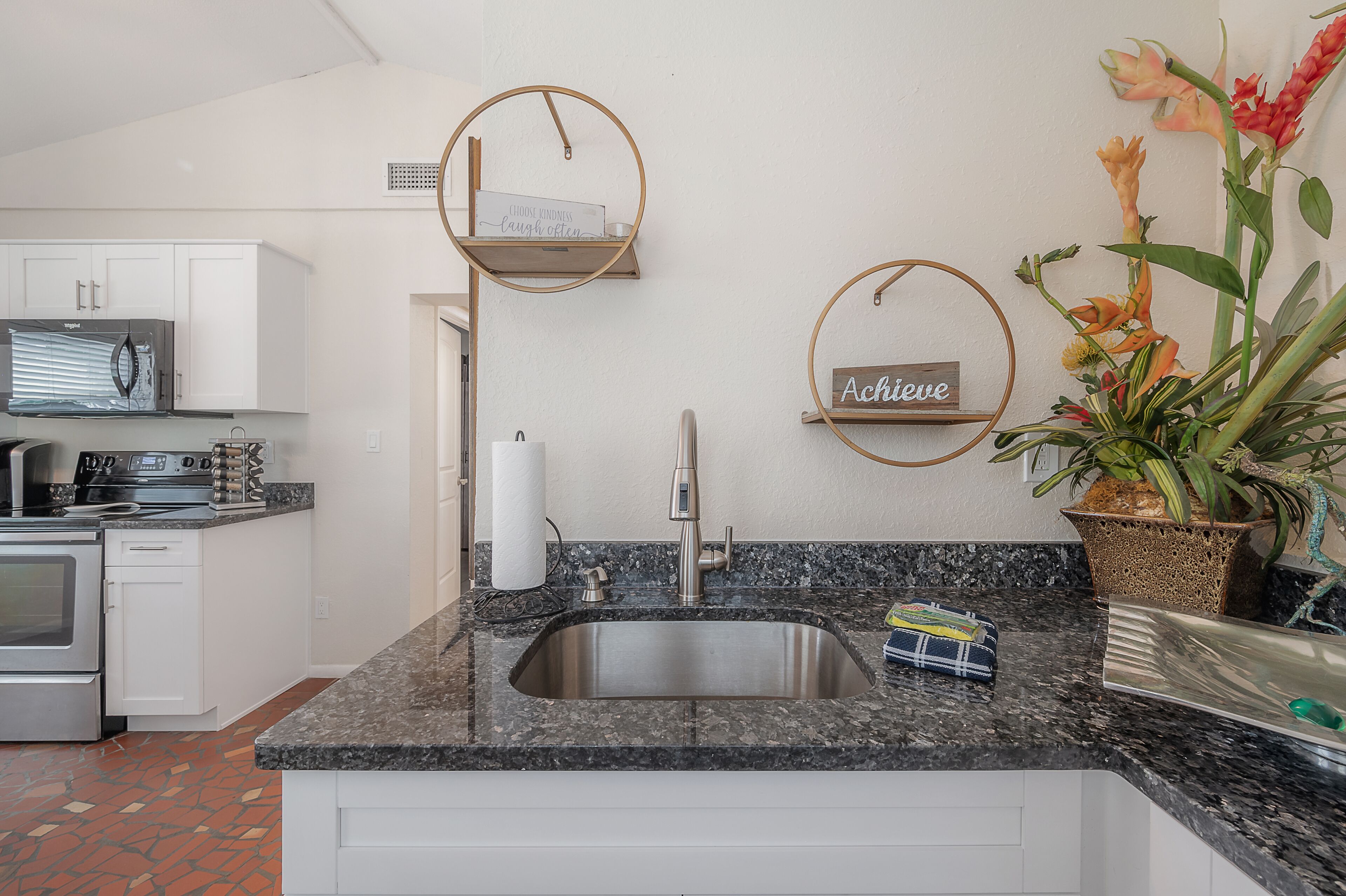 A kitchen with white cabinets, a stainless steel sink, a microwave, and a stove. There are two wall-mounted shelves with decorative items.