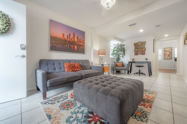 Ein modernes Wohnzimmer mit grauem Sofa, passendem Hocker, farbenfrohem Teppich, Stadtbildgemälde, Schreibtisch mit Lampe und einer Topfpflanze.