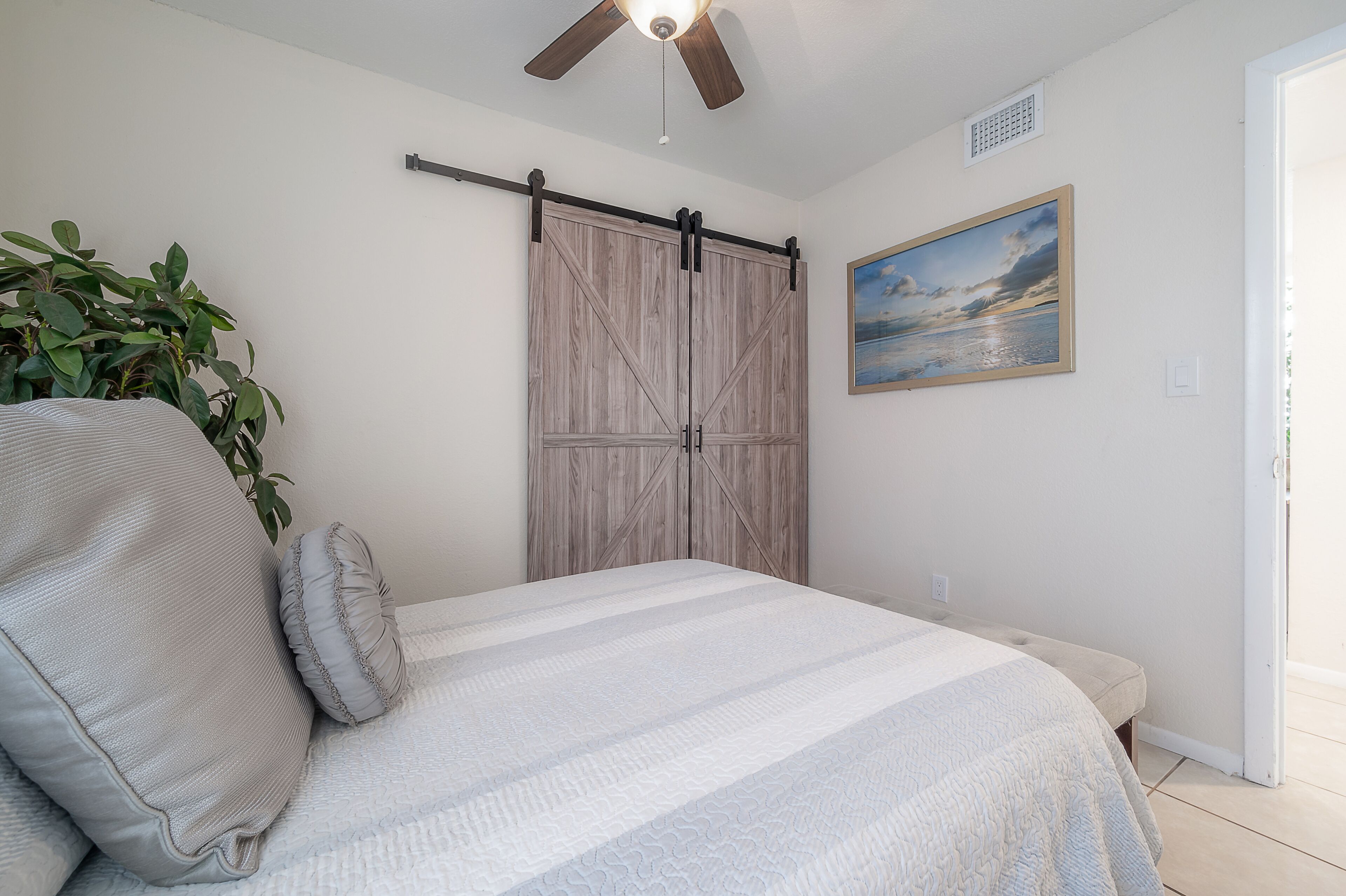 A bedroom with a bed, pillows, a plant, a painting, and a sliding door.