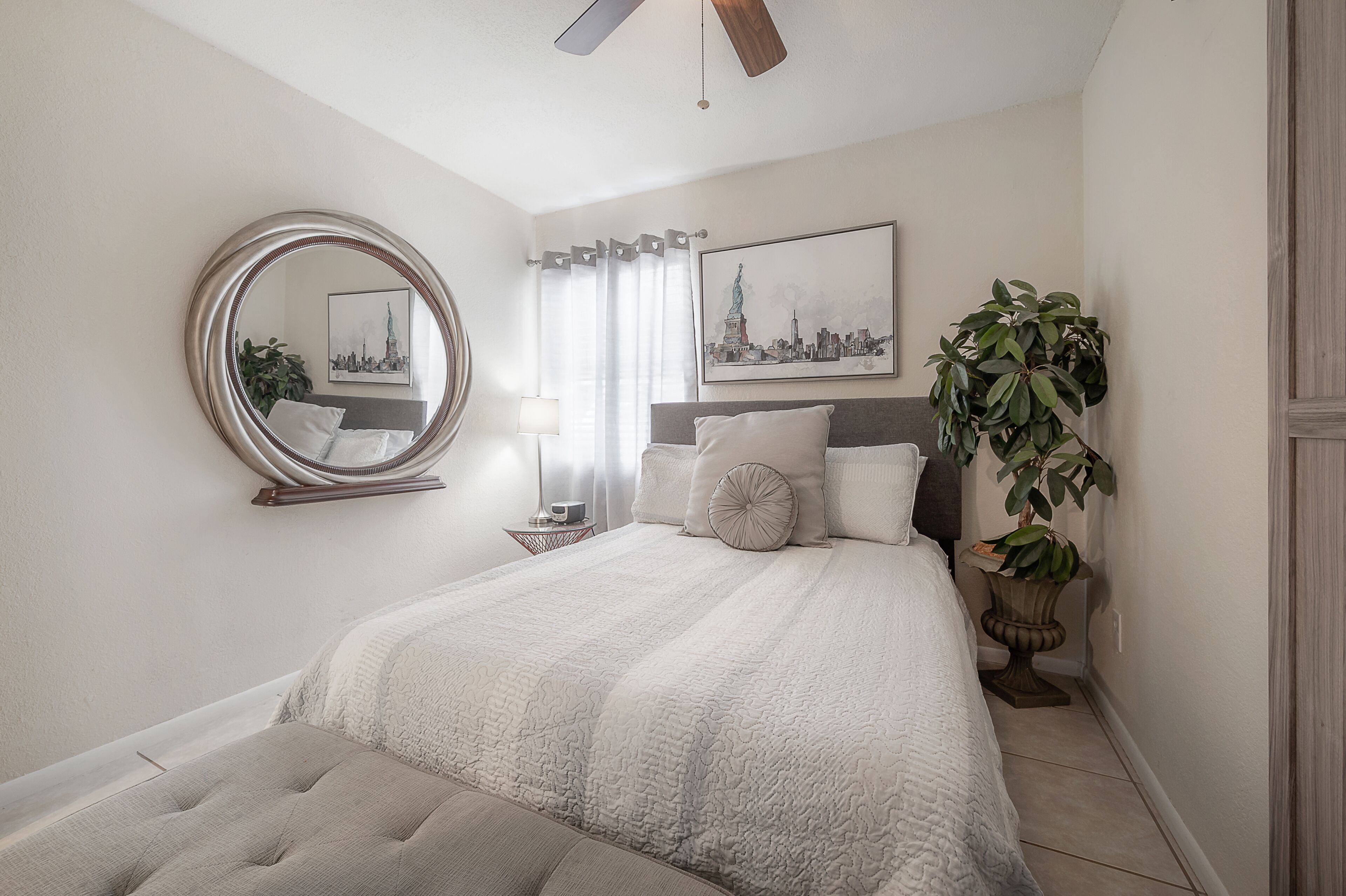 Un dormitorio con cama, espejo redondo, pintura de paisaje urbano, planta en maceta y ventilador de techo.