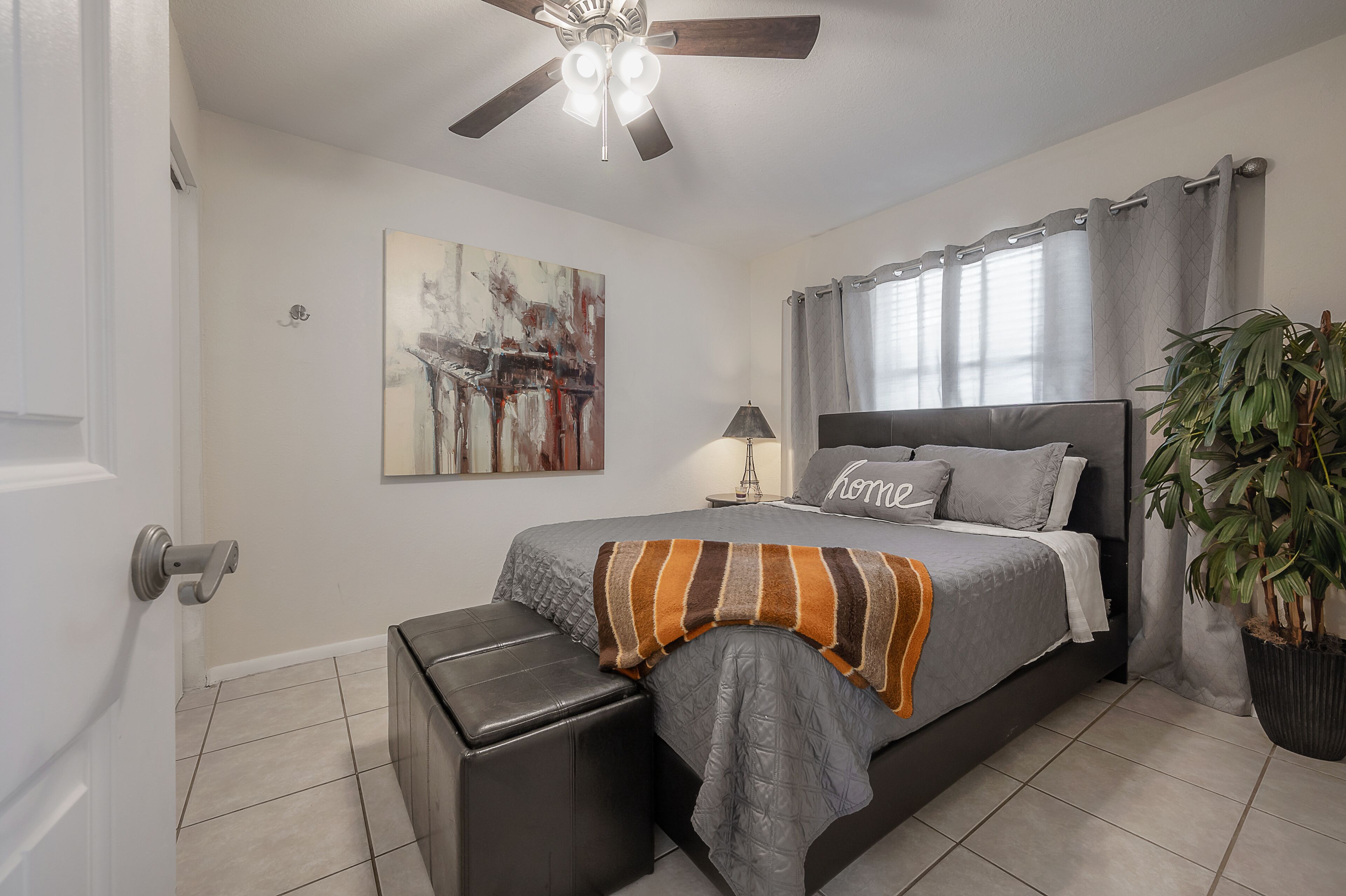 A bedroom with a bed, a ceiling fan, a nightstand, a lamp, a painting, curtains, a potted plant, and a door.
