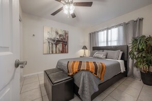 Ein Schlafzimmer mit Bett, Deckenventilator, Nachttisch, Lampe, Gemälde, Vorhängen, einer Topfpflanze und einer Tür.