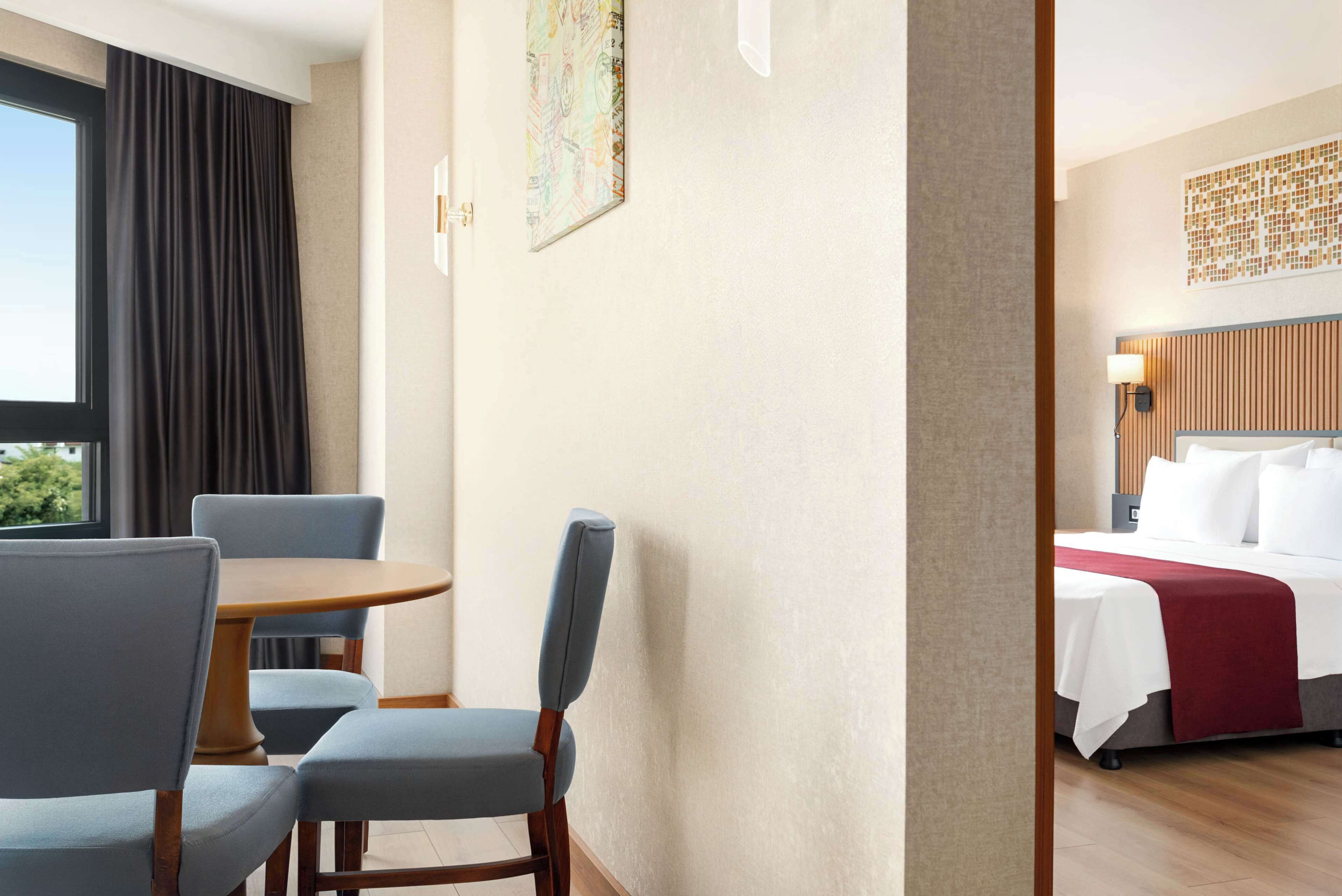 A hotel room with a bed, a dining table with chairs, a window with curtains, and a wall-mounted artwork.