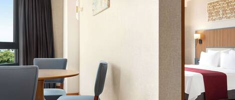 A hotel room with a bed, a dining table with chairs, a window with curtains, and a wall-mounted artwork.