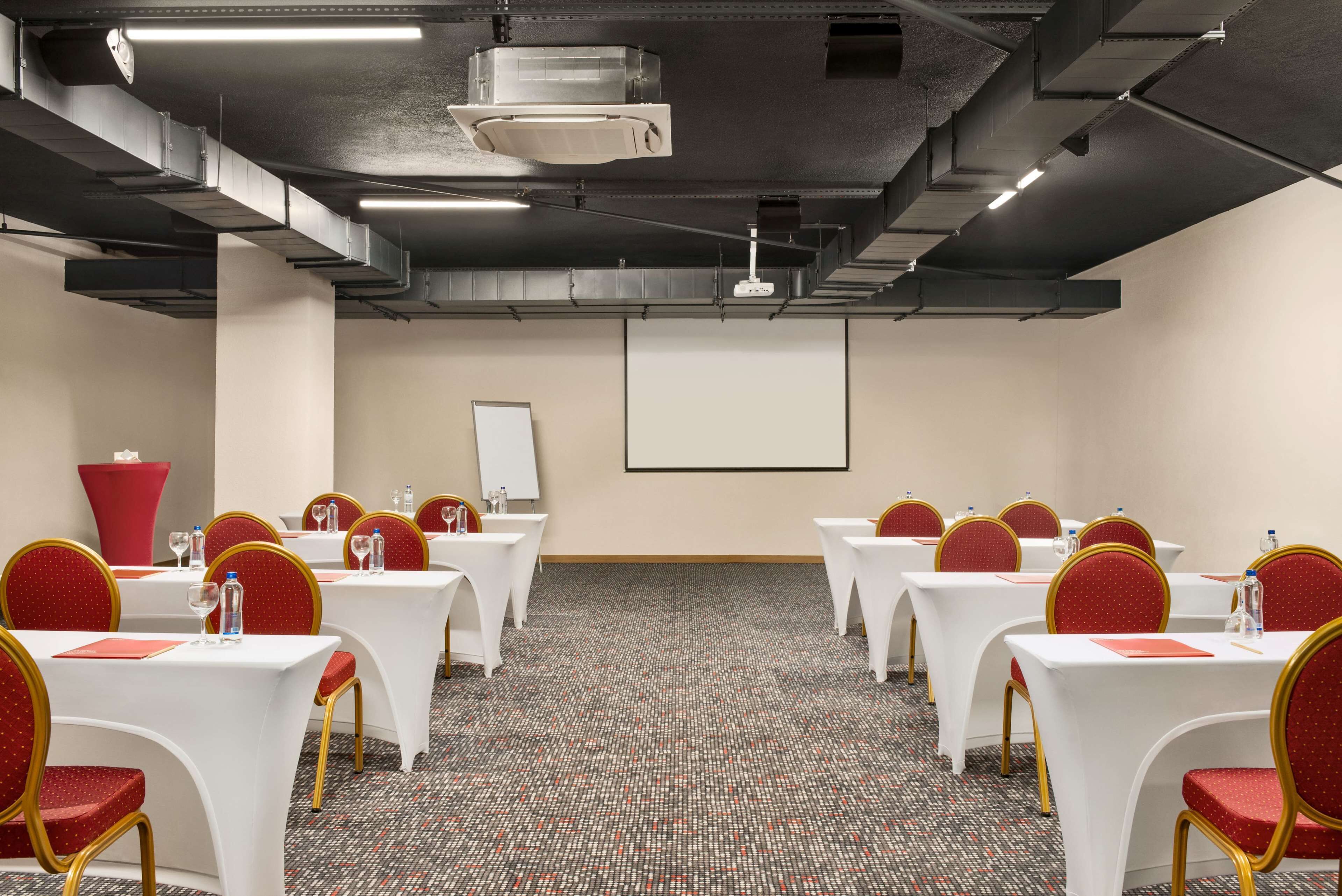 A conference room with white tables and red chairs, equipped with water bottles and glasses, and a projector screen on the wall.