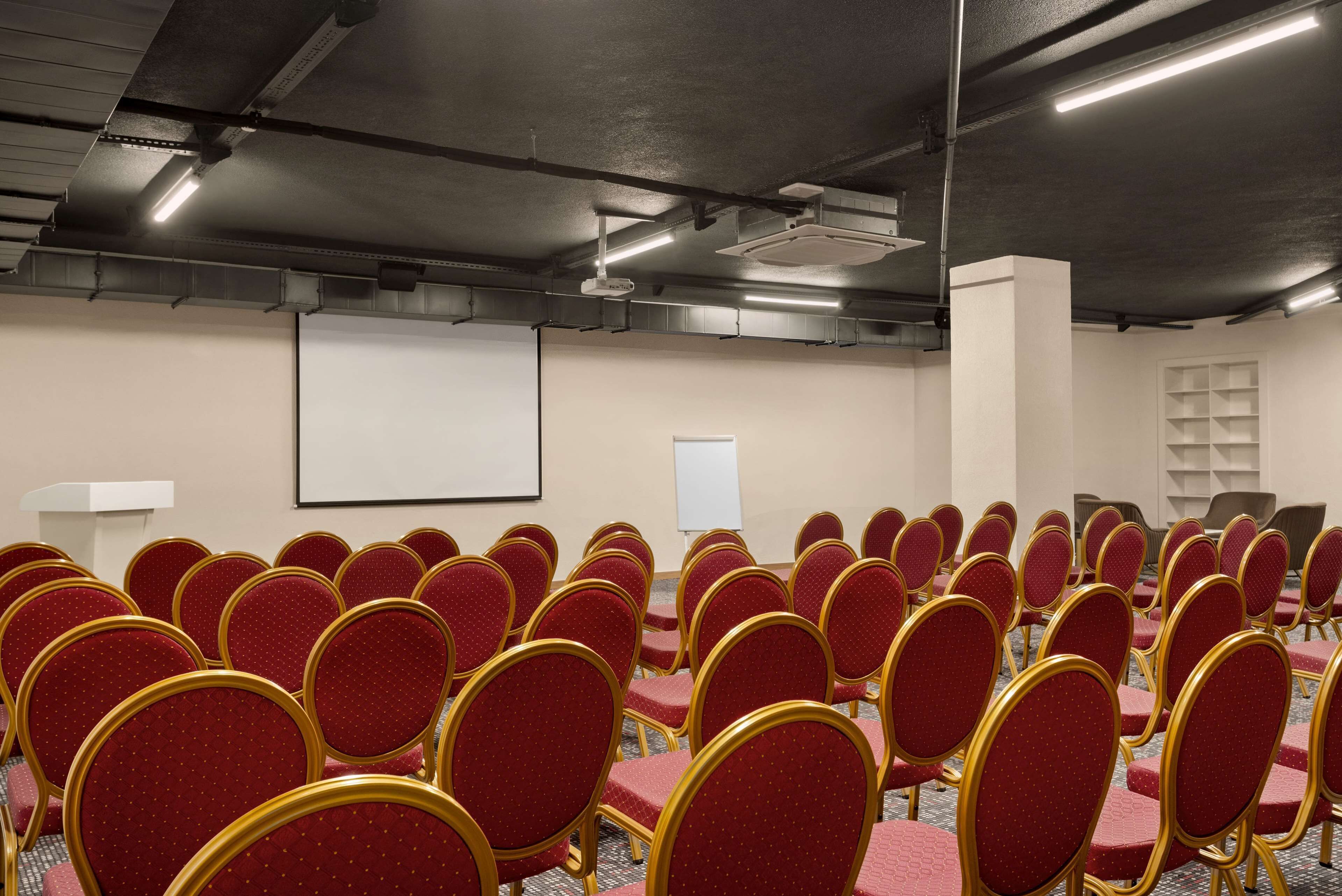 Un salón de conferencias con filas de sillas rojas y una pantalla blanca.