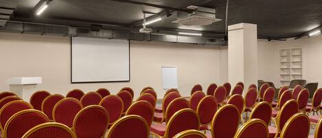 Un salón de conferencias con filas de sillas rojas y una pantalla blanca.
