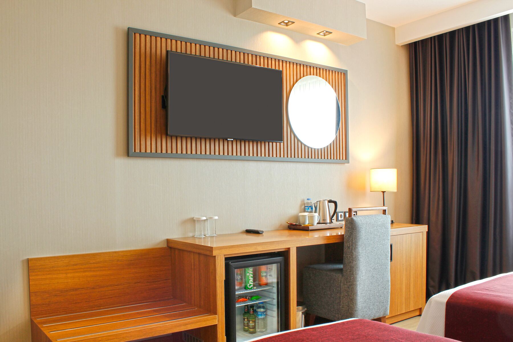 A hotel room with a wooden desk, a minibar, a television mounted above the desk, a mirror, and a bed with red bedding.