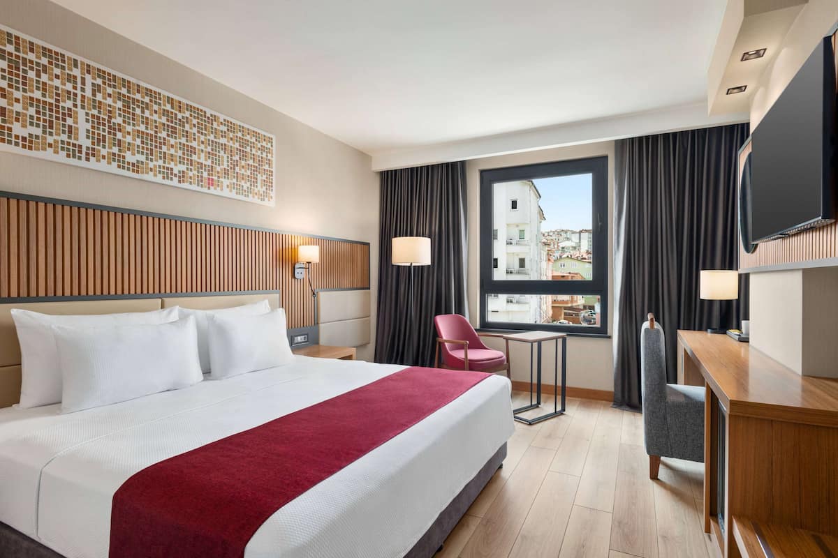 Ein Hotelzimmer mit einem großen Bett, einem Schreibtisch, einem Sessel, einem Fernseher und einem Fenster mit Blick auf die Stadt.
