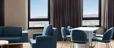 A modern living room with a sofa, armchair, and dining table with chairs. Large windows offer a city view.