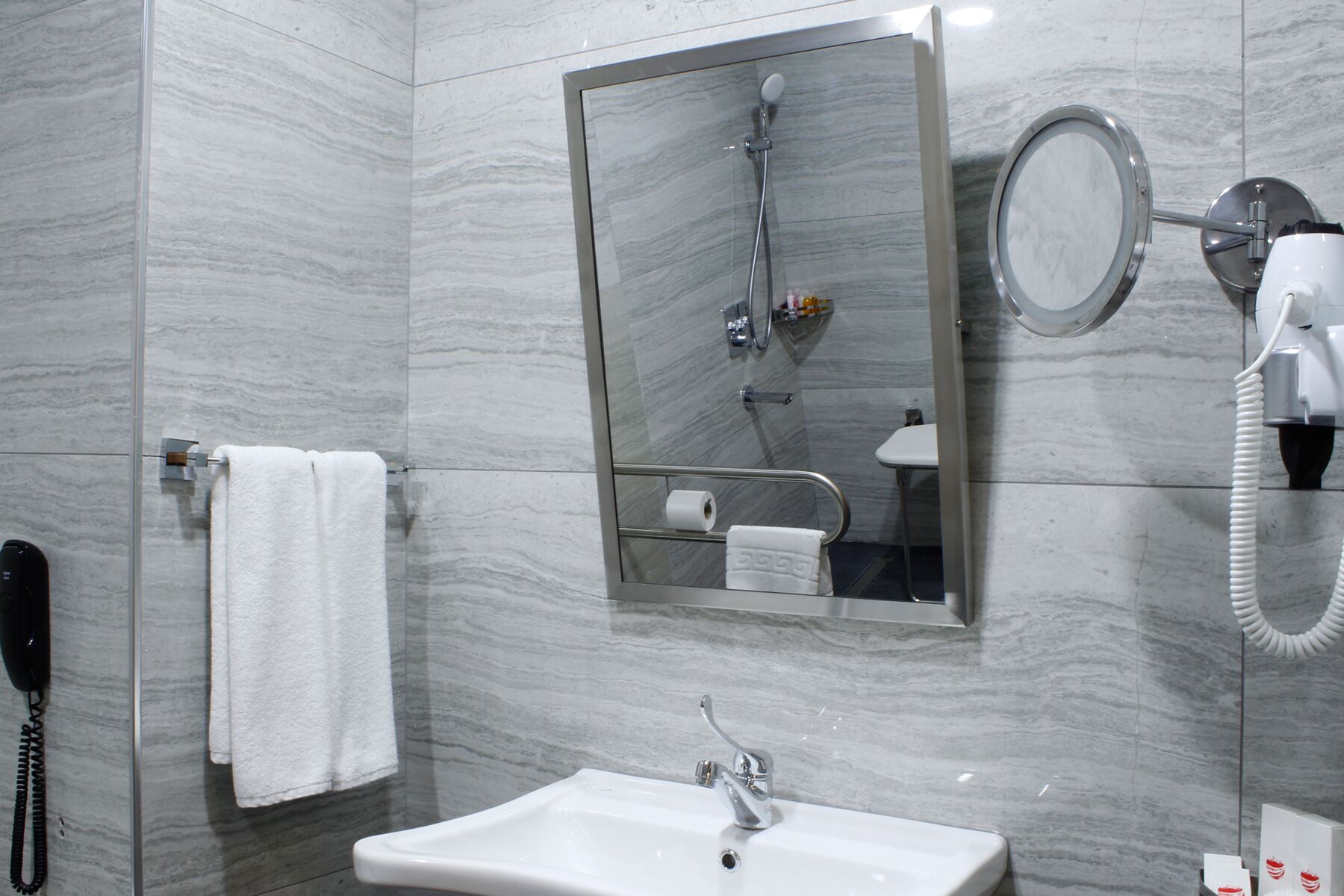 A bathroom with a marble wall, a sink, a mirror, a handheld showerhead, a towel, and a telephone.