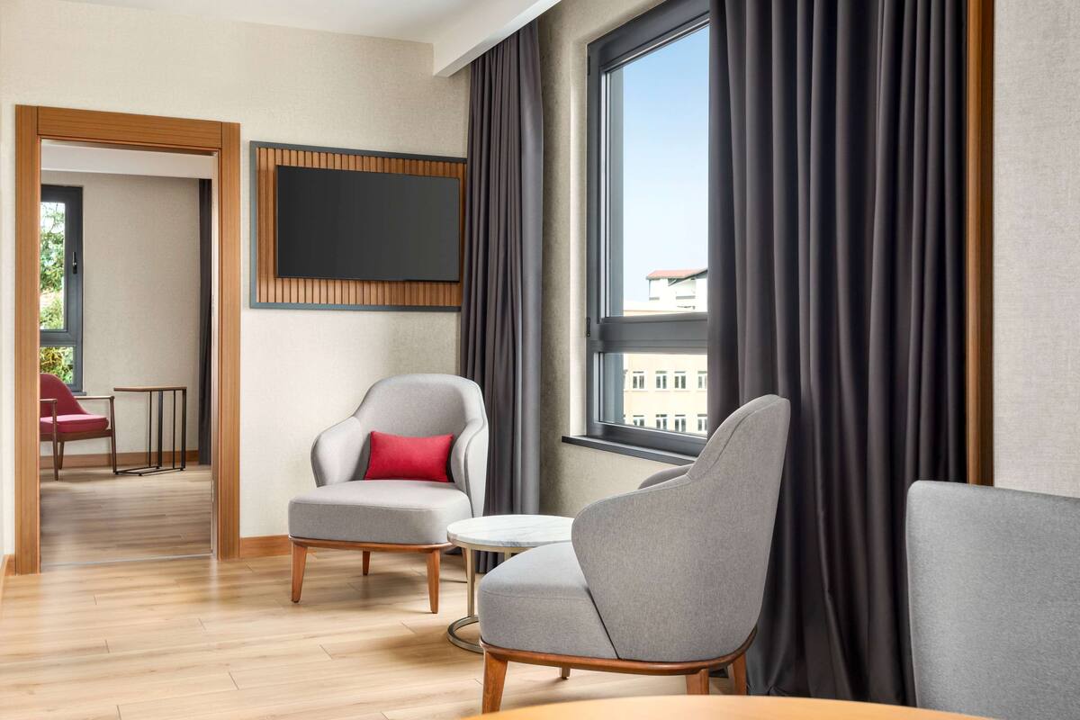 Ein modernes Hotelzimmer mit großem Fenster, grau gepolsterten Sesseln, einem kleinen runden Tisch und einem an der Wand befestigten Flachbildfernseher.