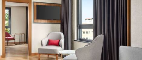 Una habitación de hotel moderna con un ventanal grande, sillones tapizados en gris, una mesita redonda y un televisor de pantalla plana montado en la pared.