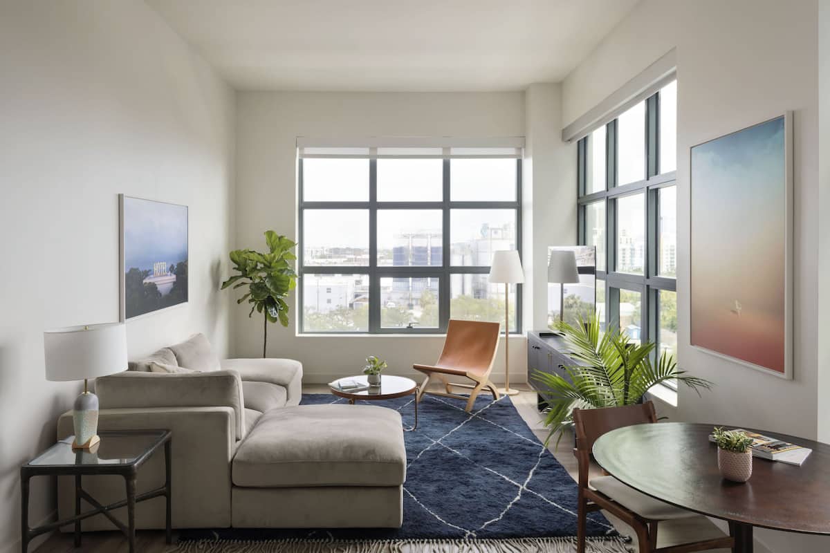 A modern hotel room with a large window, a sofa, a chair, a small table, and a dining table.