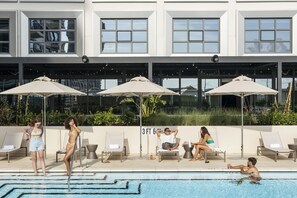 A poolside area with lounge chairs and umbrellas, a sign indicating '3 FT 6 IN' depth, and a modern building facade in the background.
