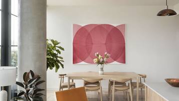 Uma sala de jantar moderna com uma mesa de madeira, cadeiras, um vaso com flores e um grande quadro abstrato na parede.