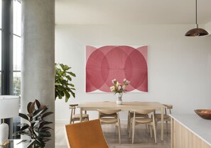 A modern dining area with a wooden table, chairs, a vase with flowers, and a large abstract painting on the wall.