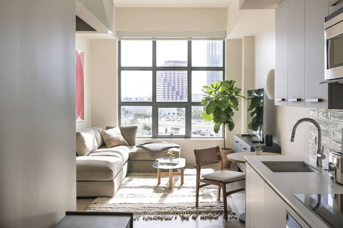 A modern living room with a large window, a sofa, a coffee table, and a kitchen area with a sink and cabinets.
