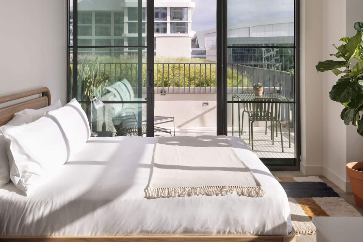 A modern bedroom with a large bed, a balcony with a view of a building, and a potted plant.