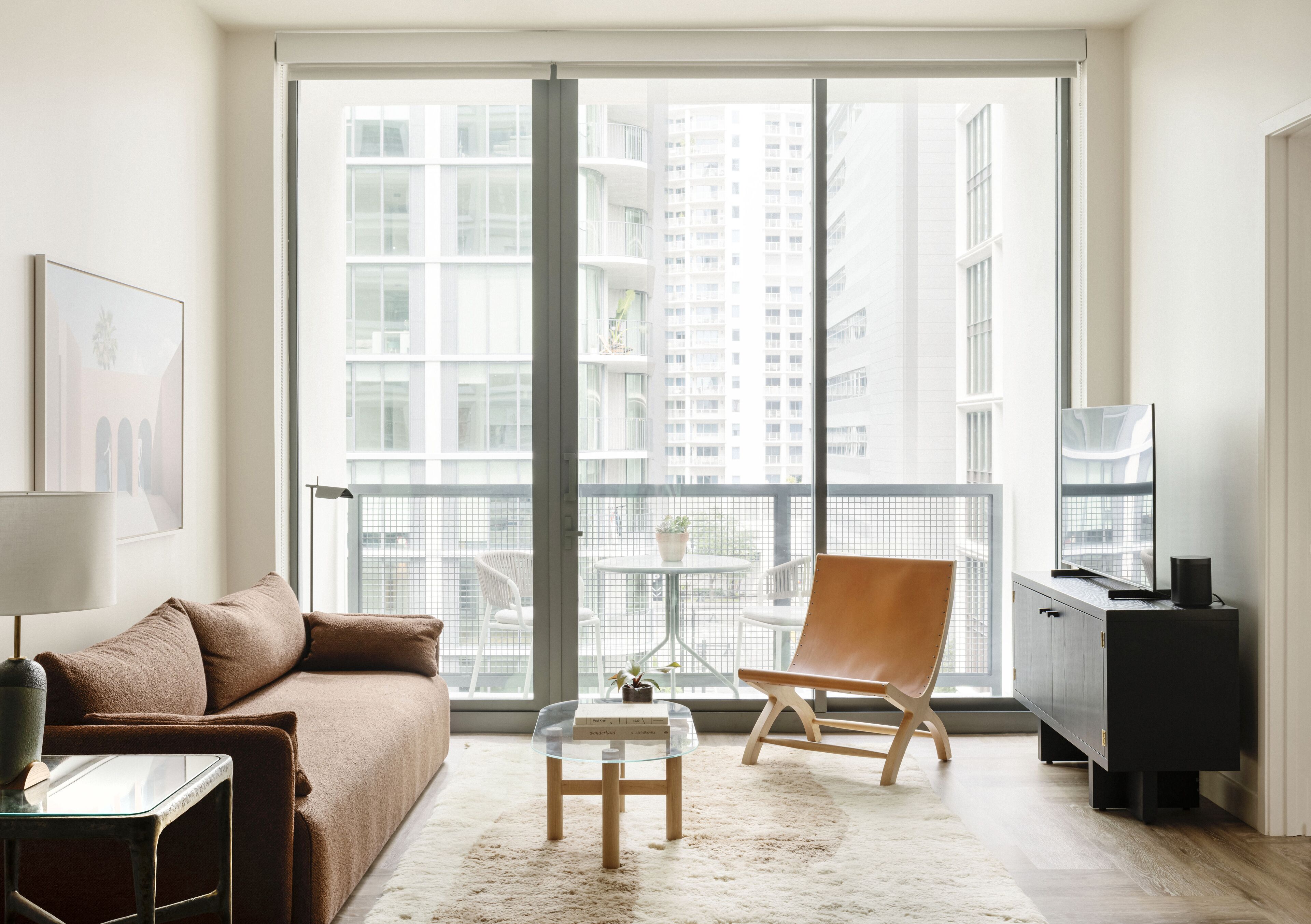 Una sala de estar moderna con un sofá, una mesa de centro de vidrio, un sillón de cuero, una mesita con una lámpara y un mueble para televisor.