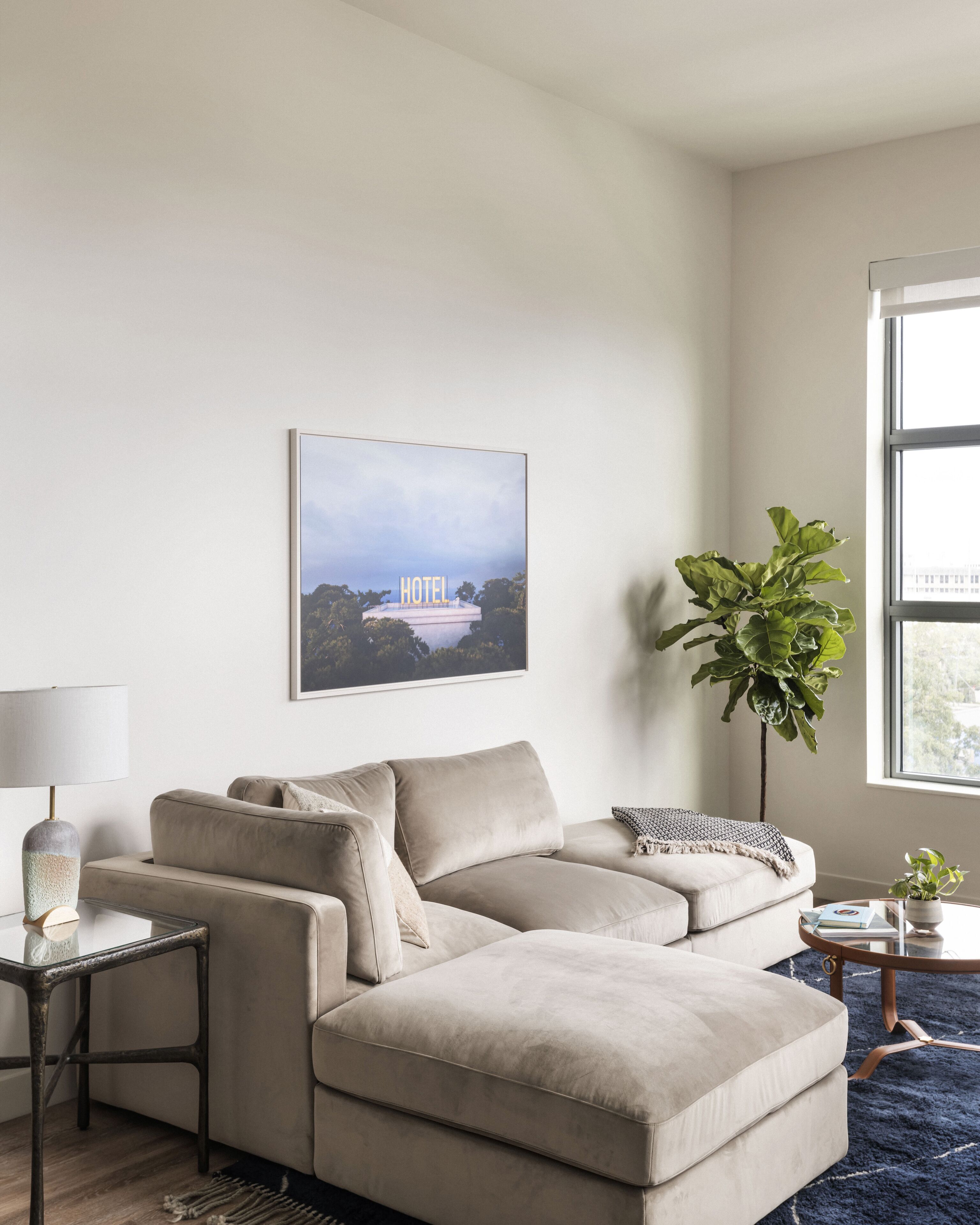 Ein modernes Wohnzimmer mit einem beigefarbenen Ecksofa, einem Glastisch, einem Beistelltisch mit Lampe, einer großen Kübelpflanze und einem gerahmten Bild eines Hotels an der Wand.