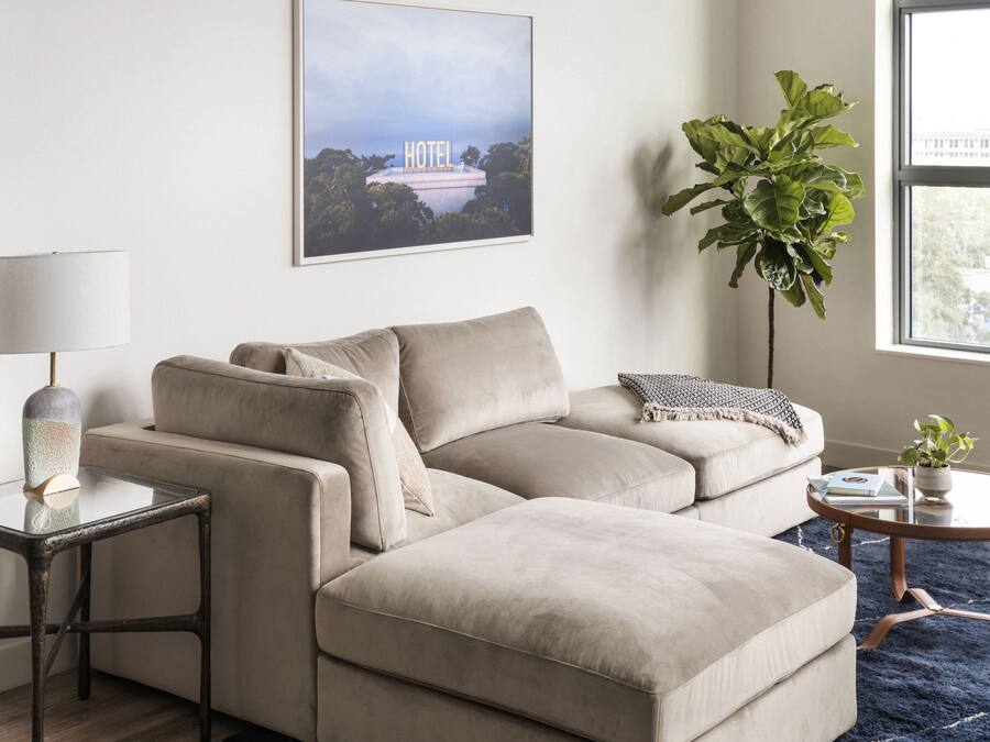 Ein modernes Wohnzimmer mit einem beigefarbenen Ecksofa, einem Glastisch, einem Beistelltisch mit Lampe, einer großen Kübelpflanze und einem gerahmten Bild eines Hotels an der Wand.