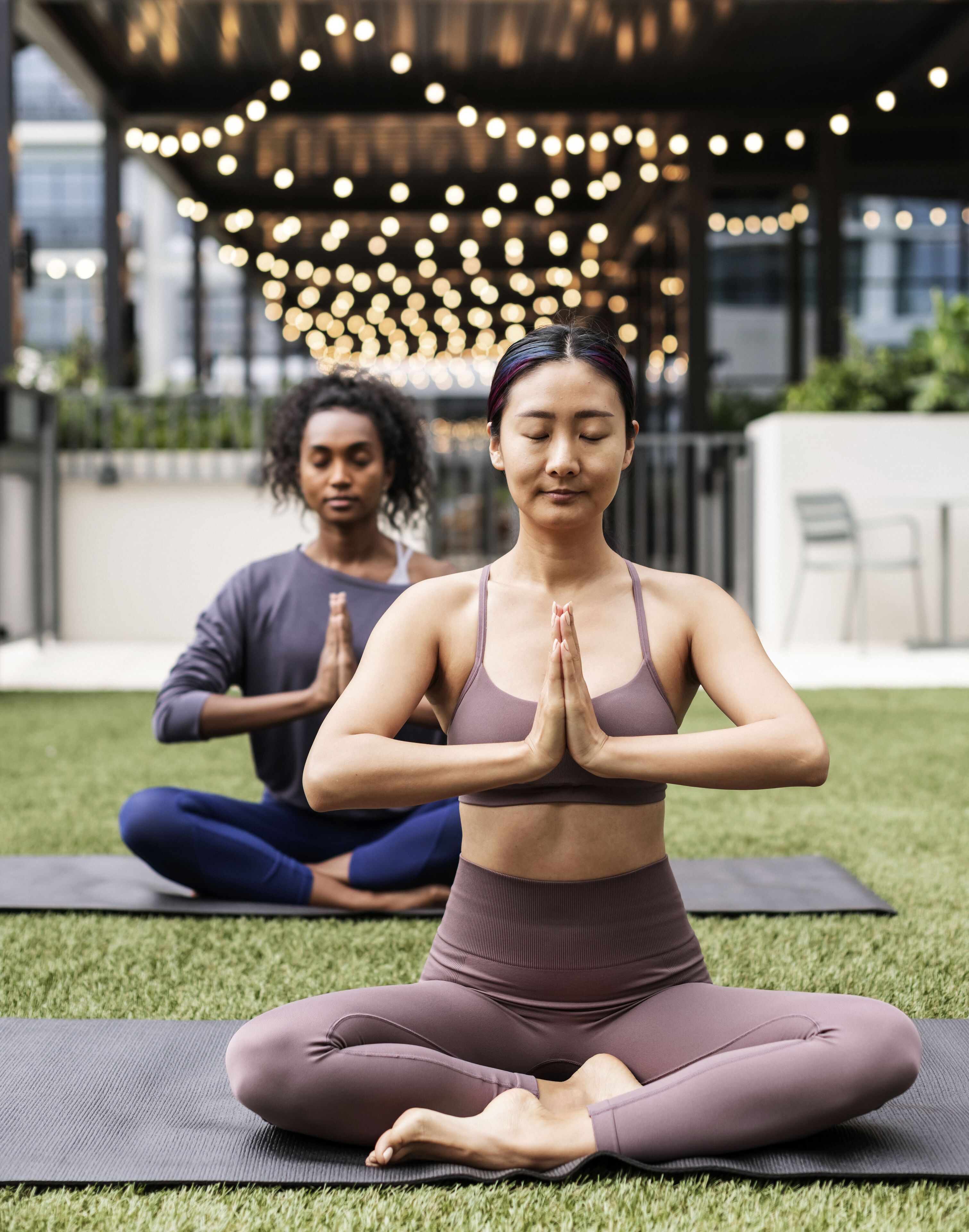 Deux personnes assises sur des tapis de yoga dans un cadre extérieur, avec en arrière-plan des guirlandes lumineuses et un bâtiment.