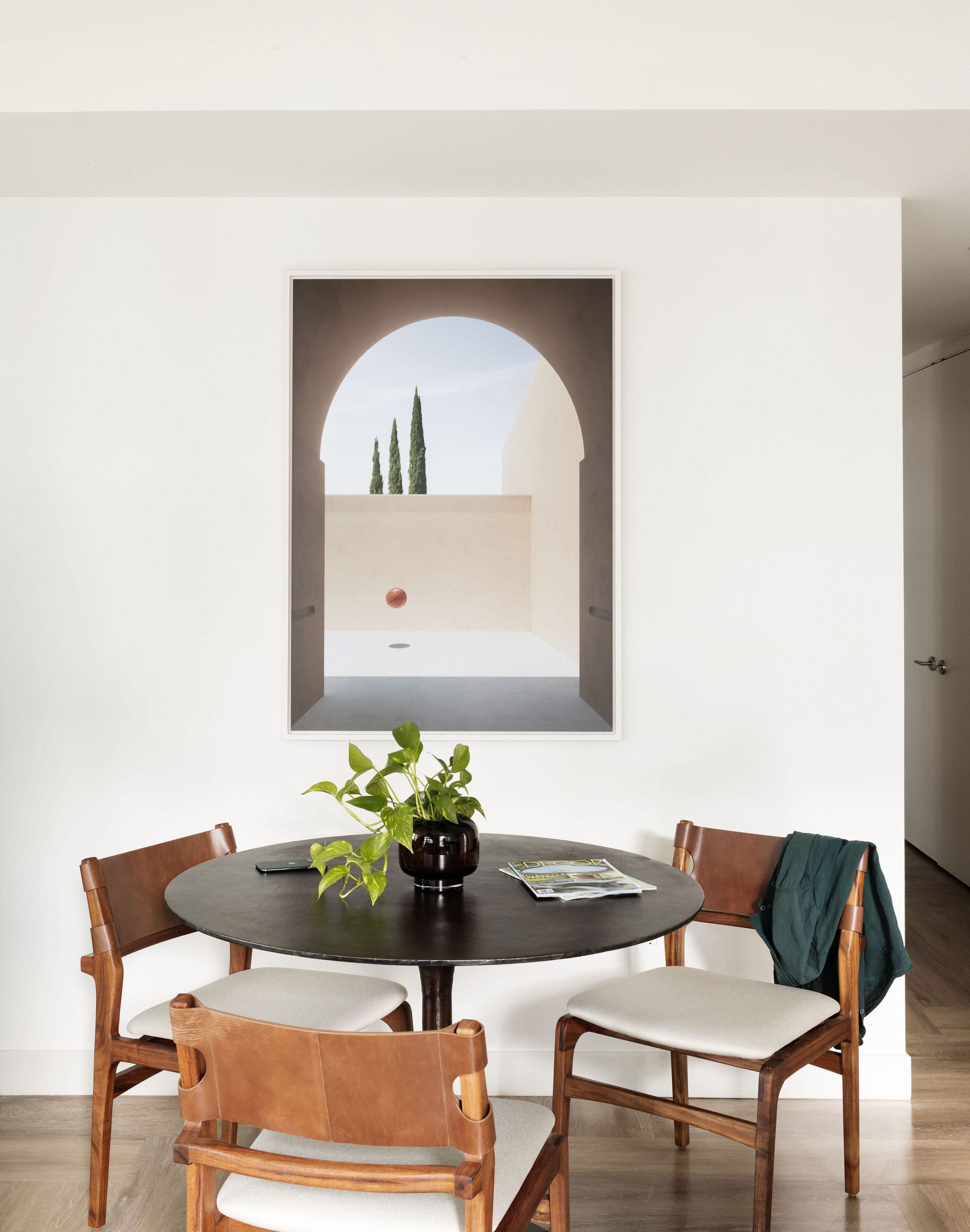 A dining area with a round table, four chairs, a potted plant, and a framed picture of an outdoor scene.