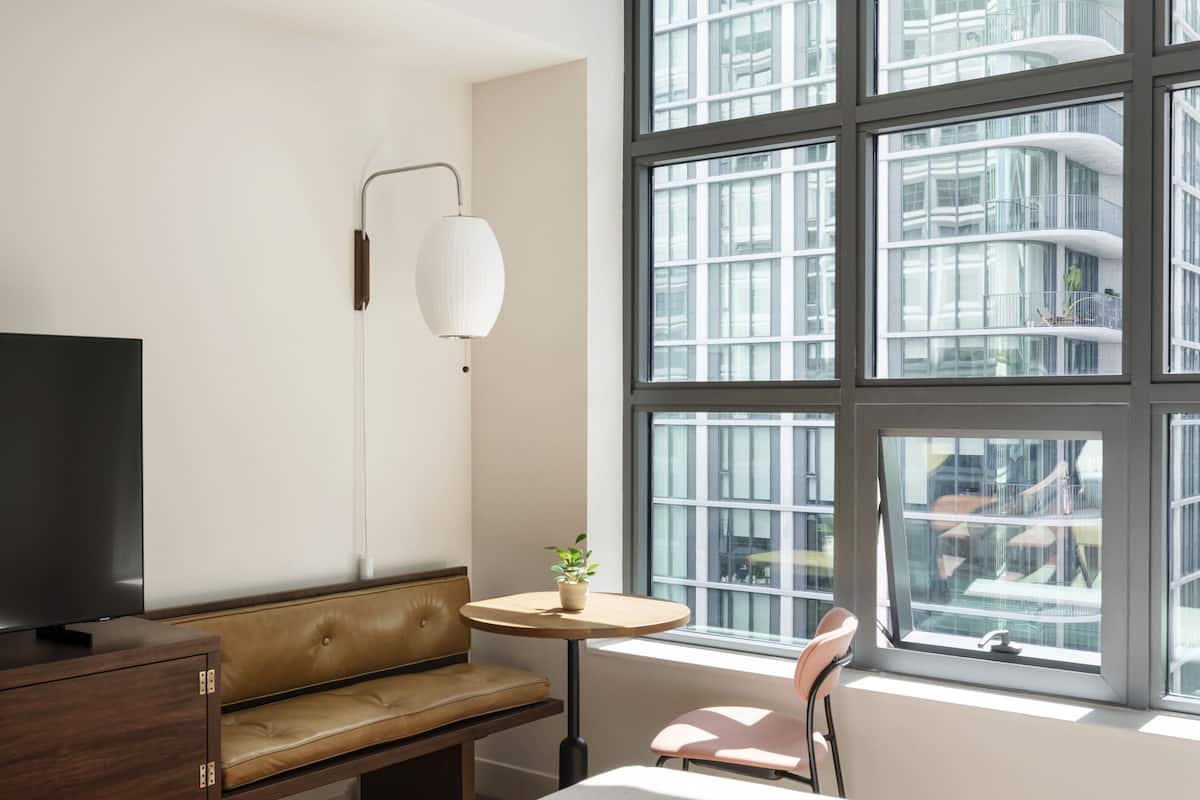 A modern hotel room with a large window, a television, a sofa, a small table, and a chair.