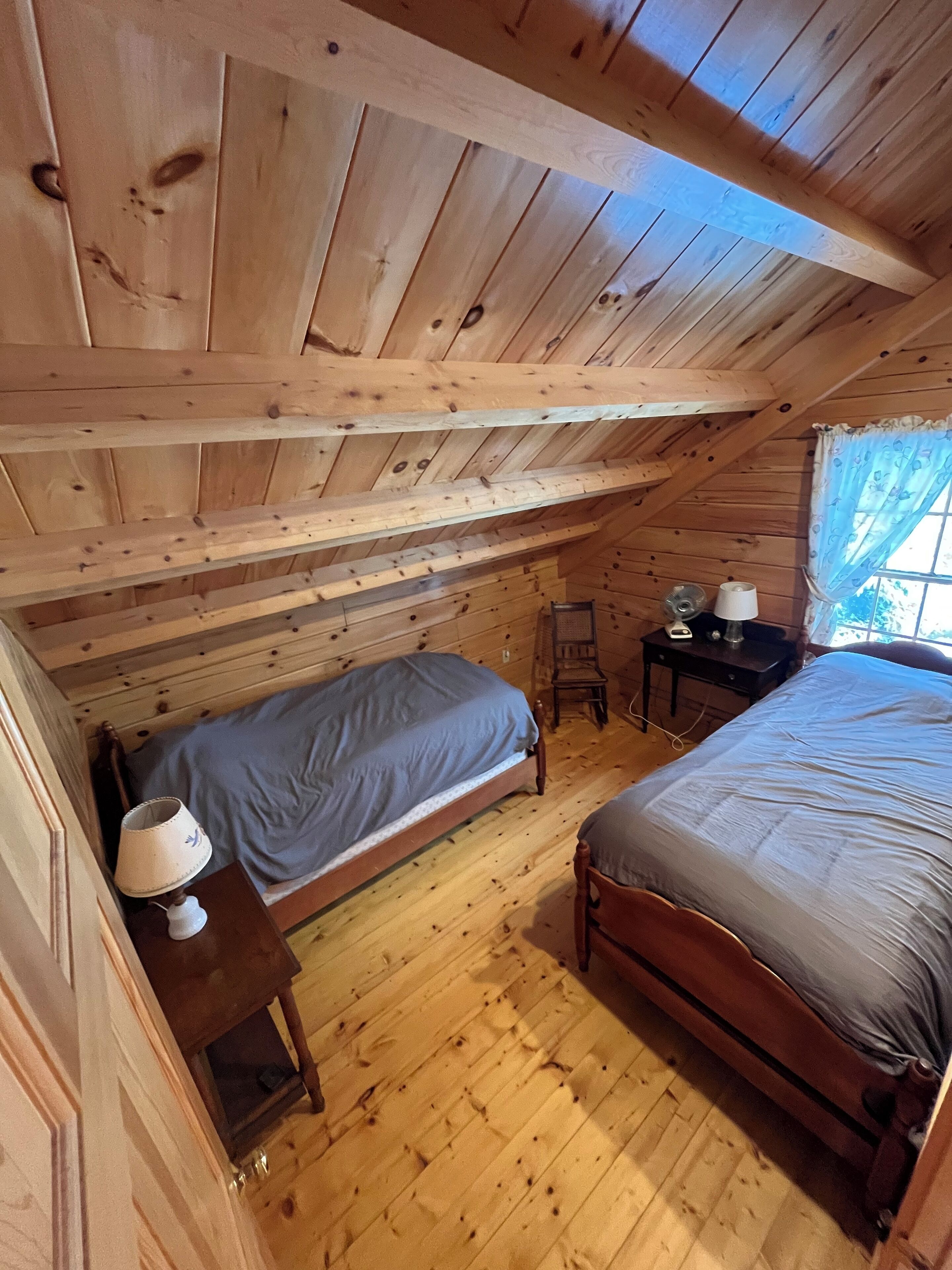 A small attic bedroom with two beds, a chair, a table, and a window with curtains.