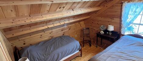 A small attic bedroom with two beds, a chair, a table, and a window with curtains.