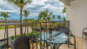 A balcony with a glass table and two wicker chairs, overlooking a beach with palm trees and a building in the background. - Ocean View, 2 Way Split AC, King Bed, Pool & WiFi (Kihei)
