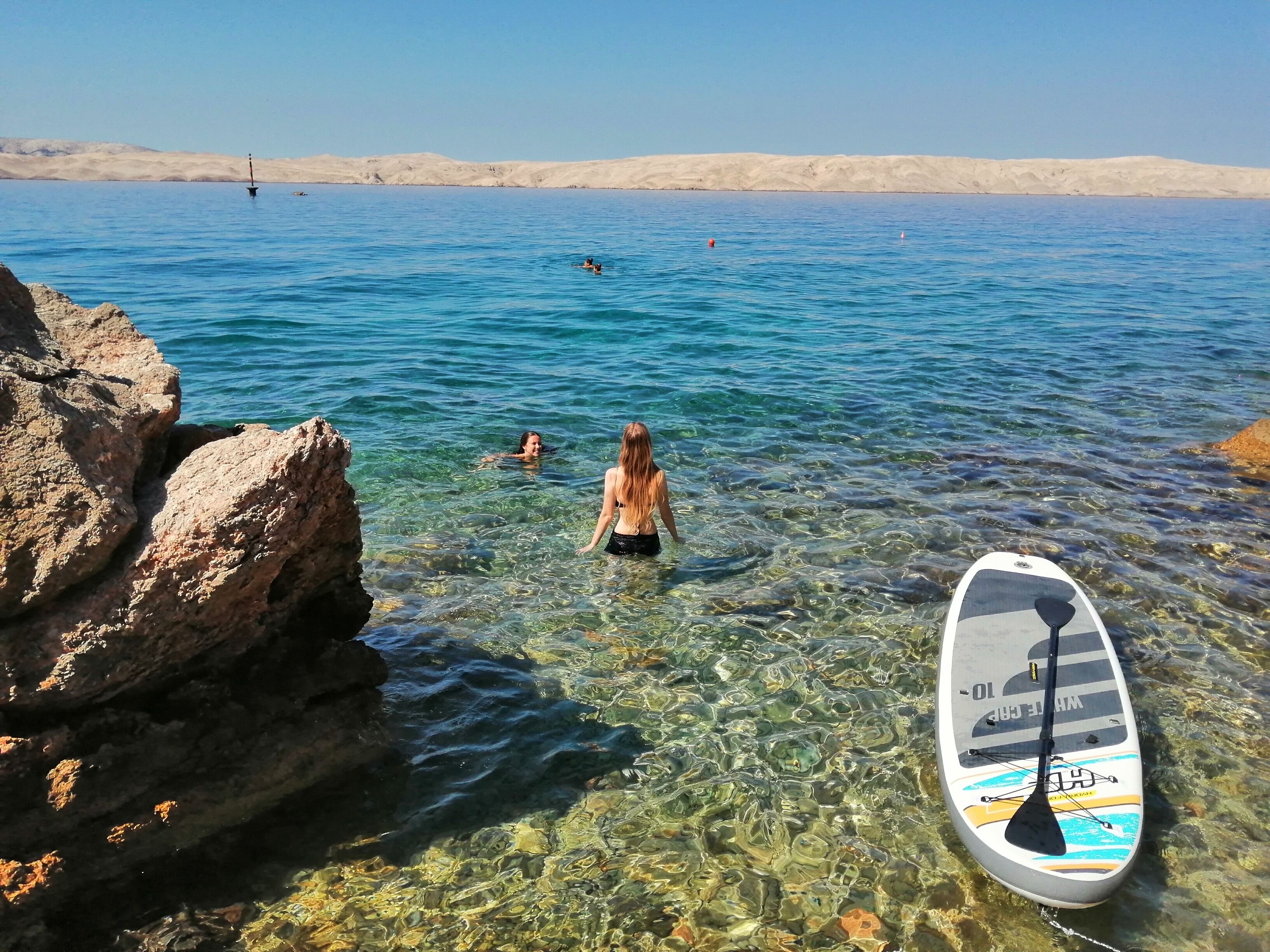 A clear lake with rocky shores, a paddleboard, and people swimming.