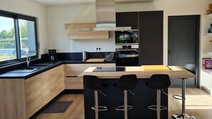 A modern kitchen with a large window, a built-in oven, and a bar area with stools. - Bright new wooden house with enclosed garden 5 minutes from the beach (Gouville-sur-Mer)