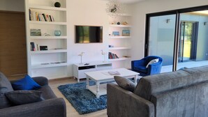 A modern living room with a flat-screen TV, white shelving, a blue chair, and a grey sofa. - Bright new wooden house with enclosed garden 5 minutes from the beach (Gouville-sur-Mer)