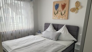 A neatly made bed with a grey headboard, white bedding, and a small bedside table with a lamp and a teddy bear. - Cozy, quiet and central, a few meters to the beach, swimming pool in the house (Norderney)