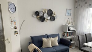 A living room with a blue sofa, a small side table with books, a bookshelf, and a clock on the wall. - Cozy, quiet and central, a few meters to the beach, swimming pool in the house (Norderney)