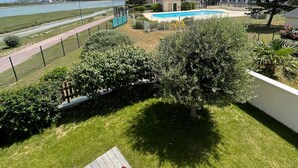 A pool area with a view of a body of water, a building with a red roof, and a wooden deck with chairs. - House with beautiful view, swimming pool, beach 5 min walk (Dives-sur-Mer)