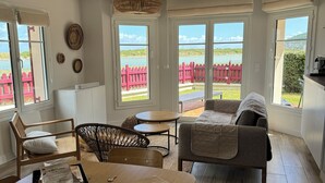 A spacious living area with a wooden dining table, a sofa, and a chair. Large windows offer views of a red fence and greenery. - House with beautiful view, swimming pool, beach 5 min walk (Dives-sur-Mer)