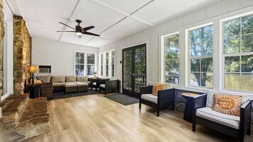 Ein geräumiges Wohnzimmer mit Kamin aus Stein, Deckenventilator und großen Fenstern mit Blick ins Freie.