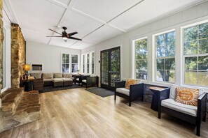 Ein geräumiges Wohnzimmer mit Kamin aus Stein, Deckenventilator und großen Fenstern mit Blick ins Freie.