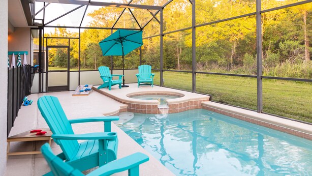 Piscina con sillas turquesa y un jacuzzi, rodeada de paredes de vidrio y vista a árboles otoñales.