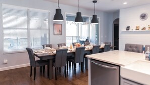 Un comedor moderno con una mesa de madera dispuesta para seis personas, lámparas colgantes negras y vistas al exterior a través de amplios ventanales.
