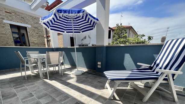 Une terrasse agrémentée d’un parasol à rayures bleues et blanches, de chaises blanches et d’une chaise longue blanche.